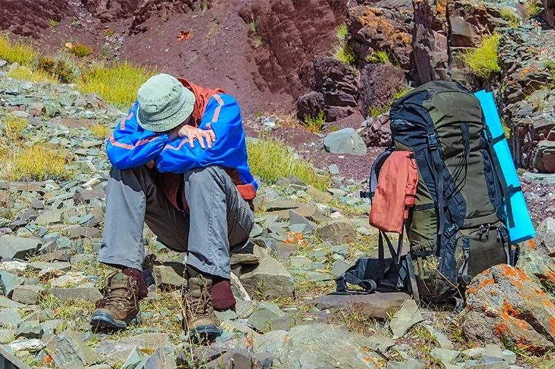 A hiker wearing a hat and blue jacket sitting on a rocky trail with his head down, resting next to a large green backpack.