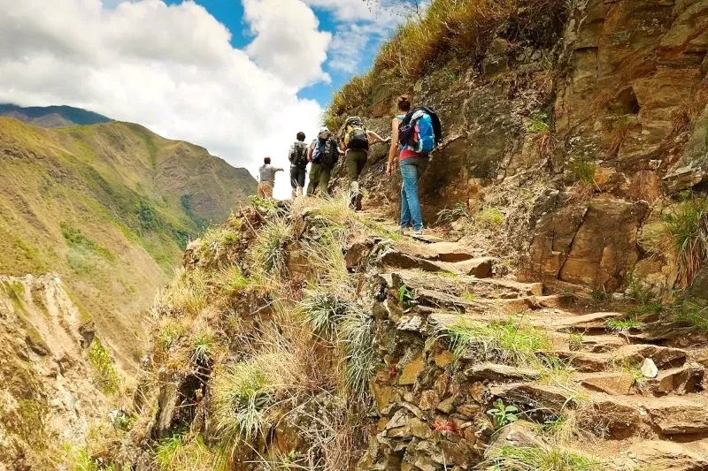 A group of hikers with backpacks walking along a narrow stone trail on a steep mountainside in the Andes.