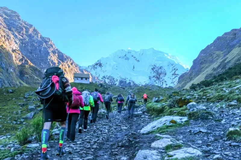A group of travelers with trekking poles climbing a rocky uphill path toward a massive glacier-covered mountain.