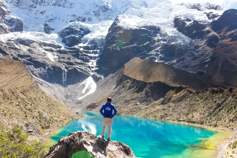 A man in a blue jacket standing on a rock looking at the turquoise Humantay Lake and a massive snow-capped glacier.