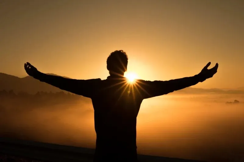 Silhouette of a person with open arms facing a golden sunrise over a misty mountain valley.