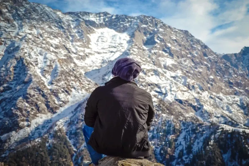 Back view of a person wearing a beanie, sitting on a rock and looking out at a massive, steep snow-covered mountain face.
