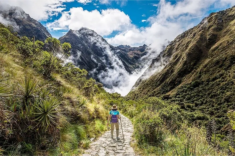 A woman hiking alone on a narrow stone-paved Inca trail surrounded by lush green mountains and misty clouds.