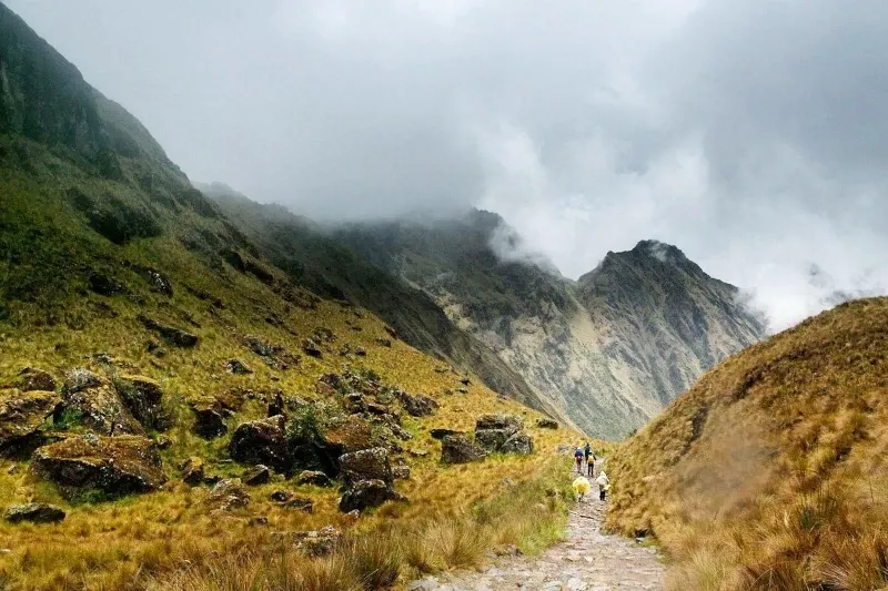 A hiker with a backpack walking on a dirt path along a mountain ridge with a snow-capped peak in the distance.