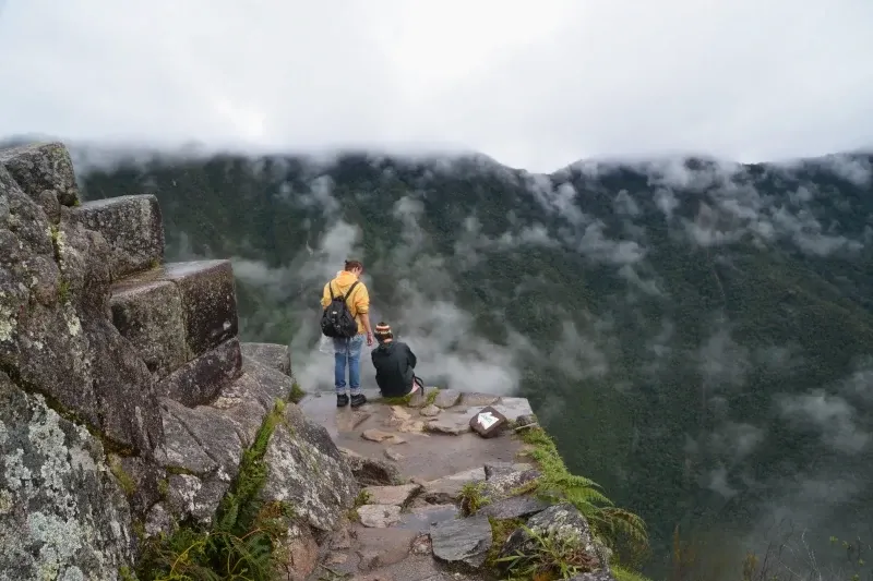 Two hikers standing on a stone ledge at the top of Huayna Picchu mountain, surrounded by mist and clouds.