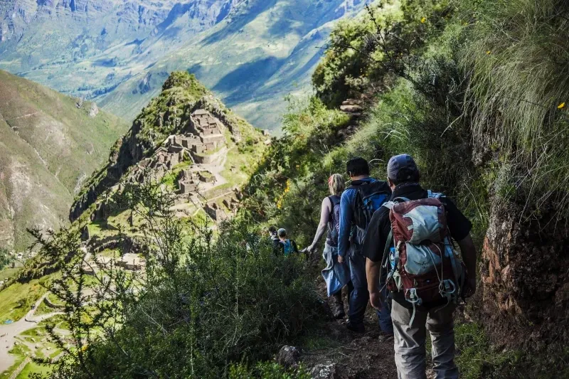 Hiking near archaeological sites in Cusco A group of hikers with backpacks descending a stone path on the Inca Trail with ancient mountain ruins in the background.