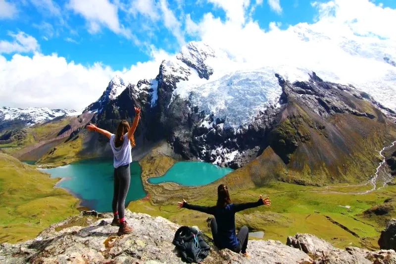 Two hikers with arms outstretched enjoying the breathtaking view of turquoise lakes and snow-covered mountains in Ausangate.