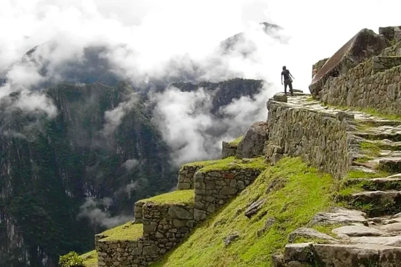 Two travelers with backpacks standing at a viewpoint overlooking a vast green mountain valley under a blue sky.