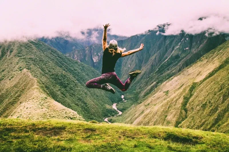 A woman jumping with joy on a mountain ridge overlooking a deep green valley and a winding river.