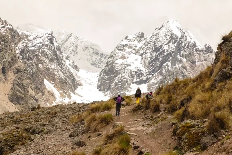 Three hikers walking on a trail towards the snow-capped Ausangate mountains in the Andes of Cusco, Peru.