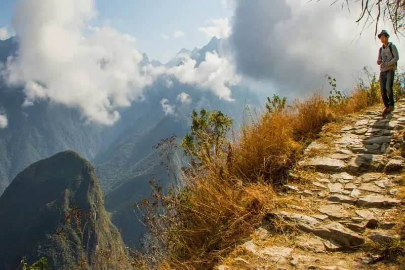 A traveler hiking on a stone path along a steep mountain ridge overlooking the lush green valleys near Machu Picchu.