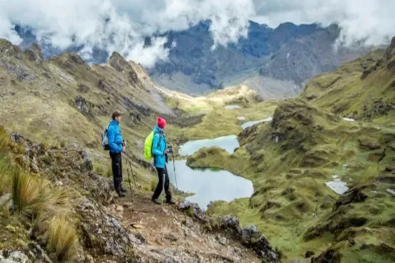Two hikers overlooking a series of bright turquoise high-altitude lakes in a green Andean valley.