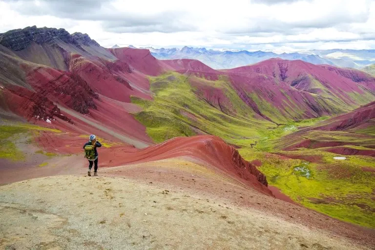 A female hiker with a green backpack walking through the stunning red and green hills of the Red Valley near Cusco.