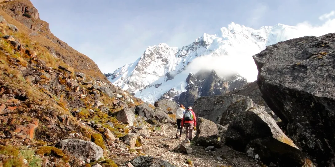 Two hikers walking on a steep, rocky mountain path toward the snowy peaks of the Peruvian Andes.