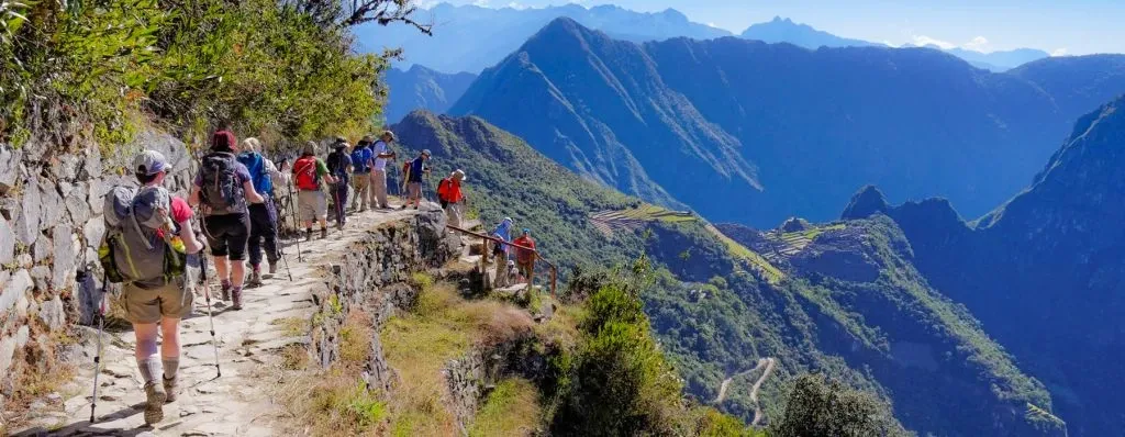 A group of trekkers with hiking poles walking along an ancient stone path on the Inca Trail with mountain views.