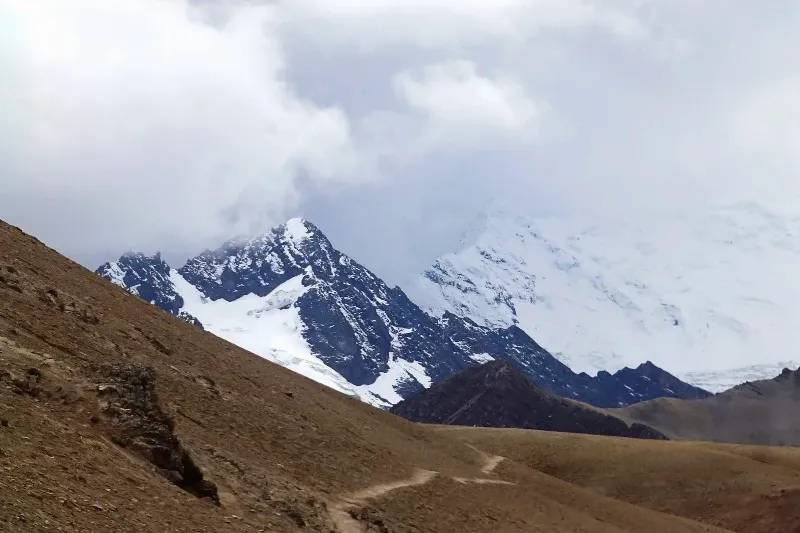A narrow hiking path winding through the brown Andean highlands toward cloudy, snow-capped mountains.