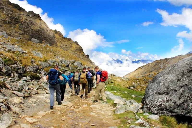 Trekkers walking on a narrow path through a valley with dramatic misty mountains and a cloudy sky.