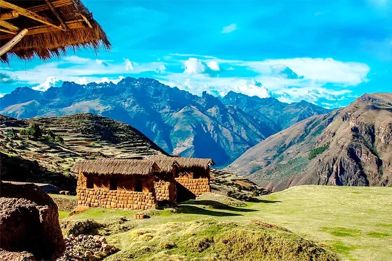 Small stone houses with thatched roofs situated on a grassy plateau with a stunning backdrop of the snow-capped Andes mountains.