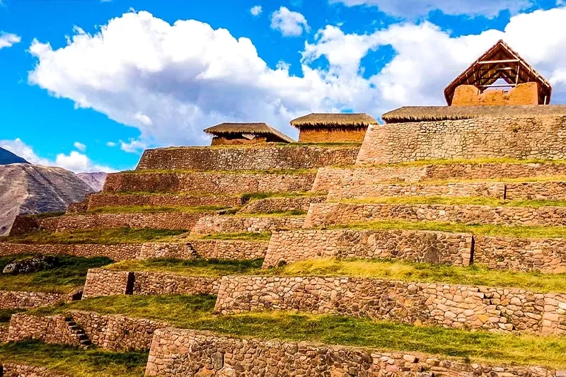 Well-preserved Incan stone buildings with thatched roofs built on a series of steep agricultural terraces under a bright blue sky.