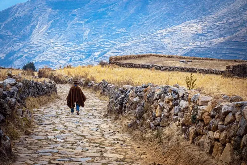 A local man wearing a traditional brown poncho walking along an ancient stone Inca path with mountain terraces in the distance.