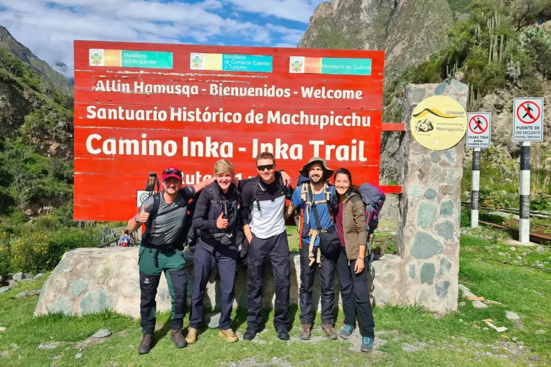 A group of five travelers posing in front of the official "Camino Inka - Inka Trail" sign at the start of the trek in Peru.