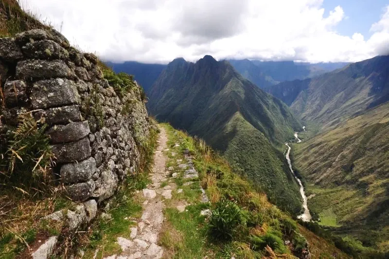 A narrow stone trail along a mountain ridge overlooking the Urubamba River winding through the valley below.
