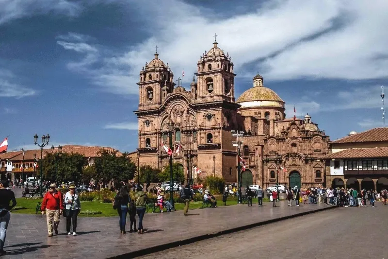 The ornate facade of La Compañia de Jesús Church in Cusco's Main Square with tourists walking by on a sunny day.
