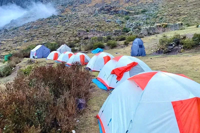 A row of white and orange camping tents set up in a green Andean valley with mountain ruins in the background.