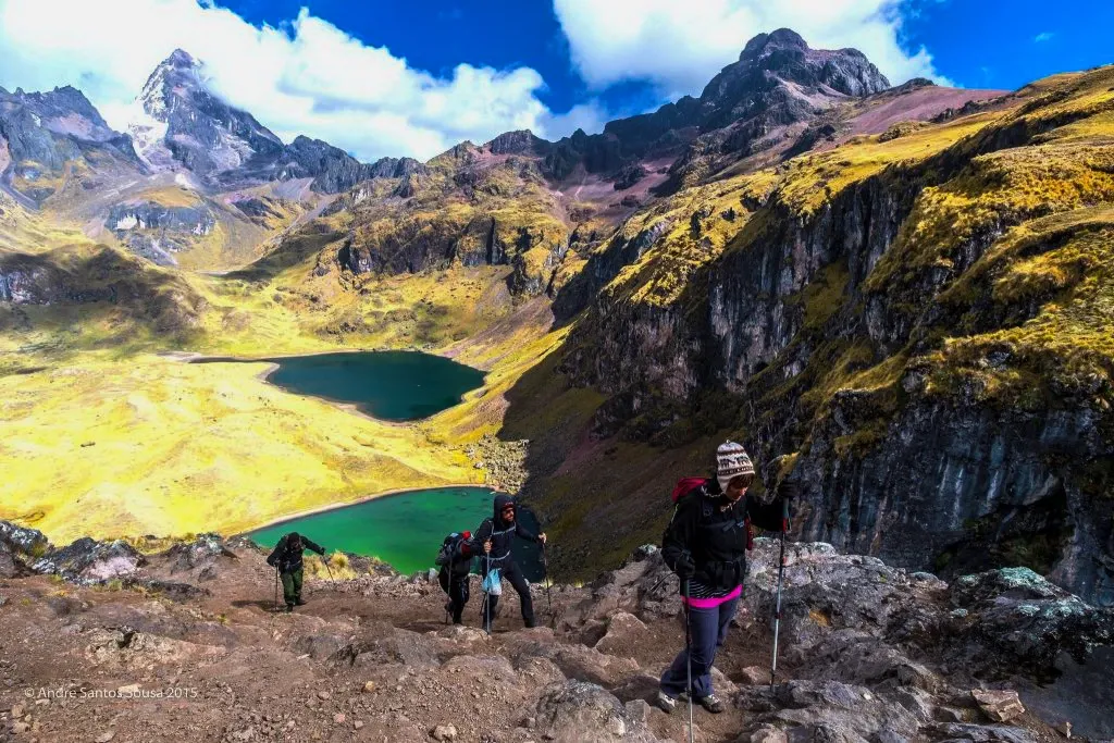 Hikers descending a rocky slope overlooking two vibrant mountain lakes, one green and one turquoise, in the Lares region of Cusco.