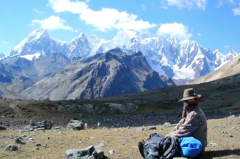 A hiker sitting on a rock resting while looking at the snow-capped peaks of the Andes during the Lares Trek.