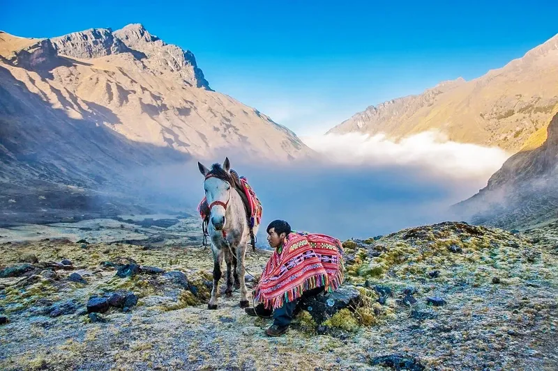 A local man wearing a traditional Peruvian poncho sitting next to a horse in a misty mountain valley.
