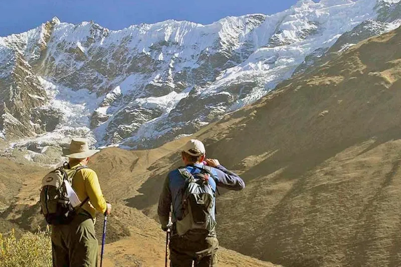 Two hikers with backpacks looking at a massive snow-capped mountain during the Lares Trek in Cusco.