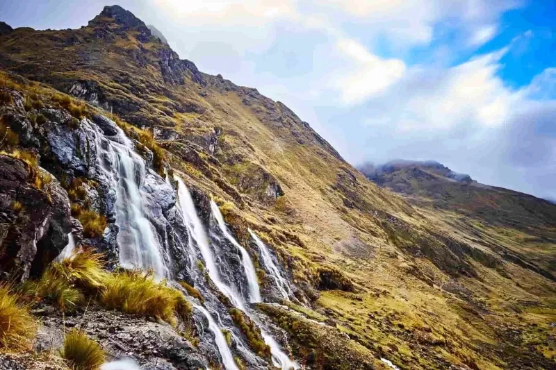 A beautiful natural waterfall flowing down a steep mountain slope under a cloudy blue sky in the Andes.