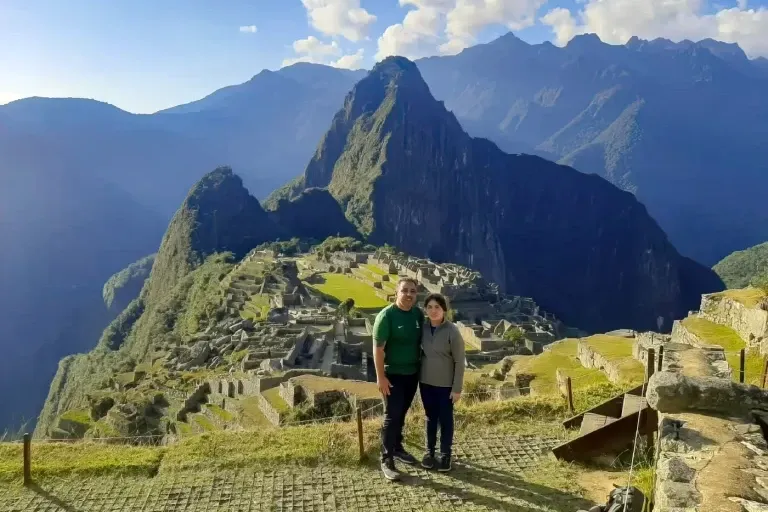 A couple posing for a photo at the classic viewpoint of Machu Picchu with Huayna Picchu mountain in the background under a clear blue sky.
