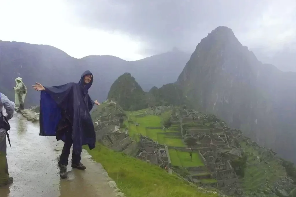 A happy traveler wearing a blue rain poncho stands on a stone path overlooking the Machu Picchu ruins during a misty rainy day.