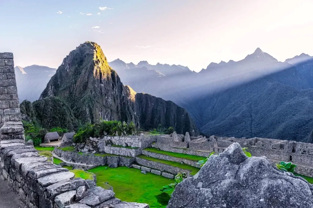 Panoramic view of the Machu Picchu stone ruins and Huayna Picchu mountain with dramatic sunbeams piercing through the Andean peaks.