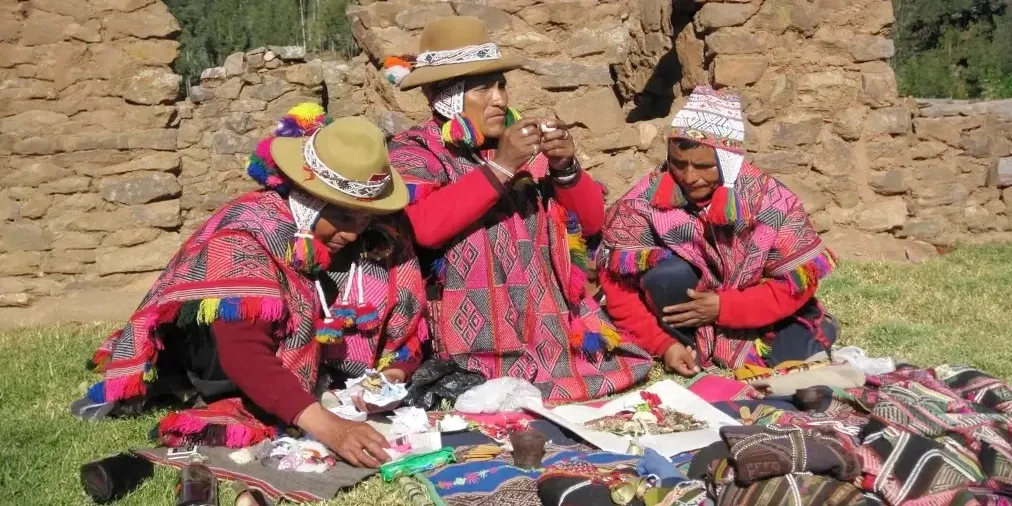 Andean shamans performing a traditional Pachamama (Mother Earth) ceremony with offerings and coca leaves in front of stone ruins.