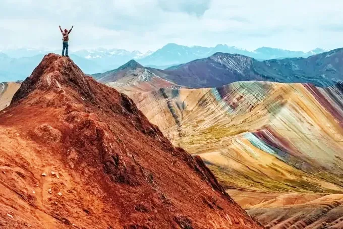A hiker standing with arms raised on a peak at Palcoyo Rainbow Mountain with snow-capped mountains in the background.