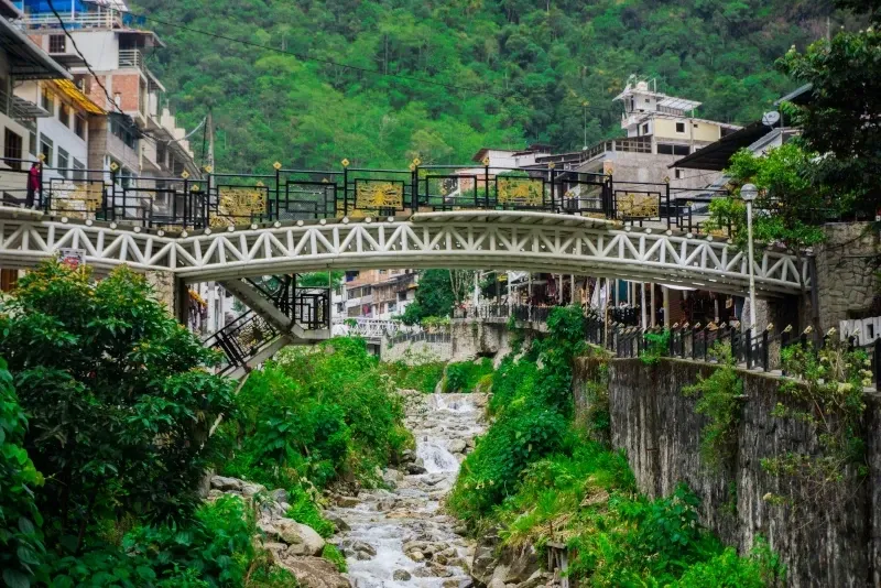 A white decorative pedestrian bridge crossing over a rocky stream in the center of Aguas Calientes.