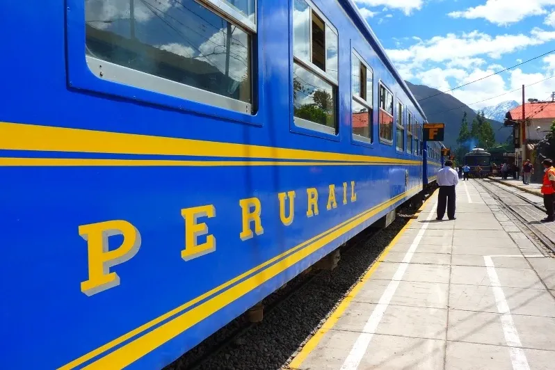 Close-up of a blue and yellow PeruRail train parked at the station under a cloudy sky.