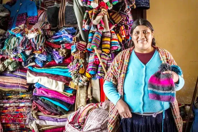 Traditional Andean textiles and handmade beanies in Cusco A smiling local woman in Cusco holding a colorful handmade wool beanie in front of a display of traditional Peruvian textiles.