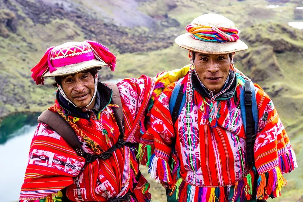 Two Quechua men wearing traditional decorated hats and red ponchos standing against a mountainous landscape with a lake.