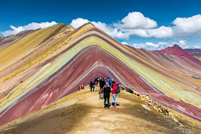 Tourists standing on the ridge of the colorful Rainbow Mountain (Vinicunca) under a bright blue sky with white clouds.