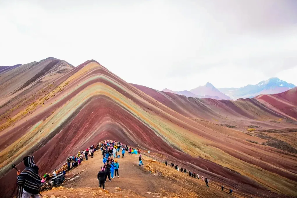 Panoramic view of Vinicunca Rainbow Mountain with tourists walking along the colorful mineral ridges under a cloudy sky.