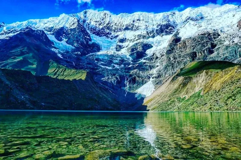 Crystal clear water of a glacial lake reflecting the towering snow-covered peaks of the Salkantay mountain range.