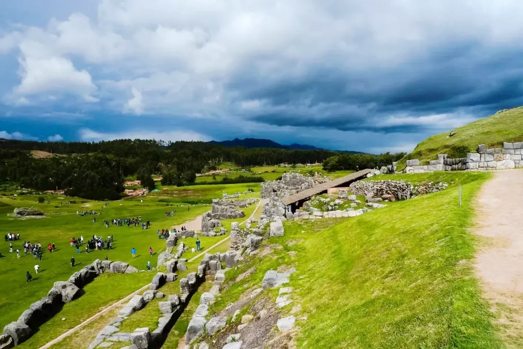 Large limestone walls of the Sacsayhuamán fortress with a vast green esplanade under a dramatic sky in Cusco.