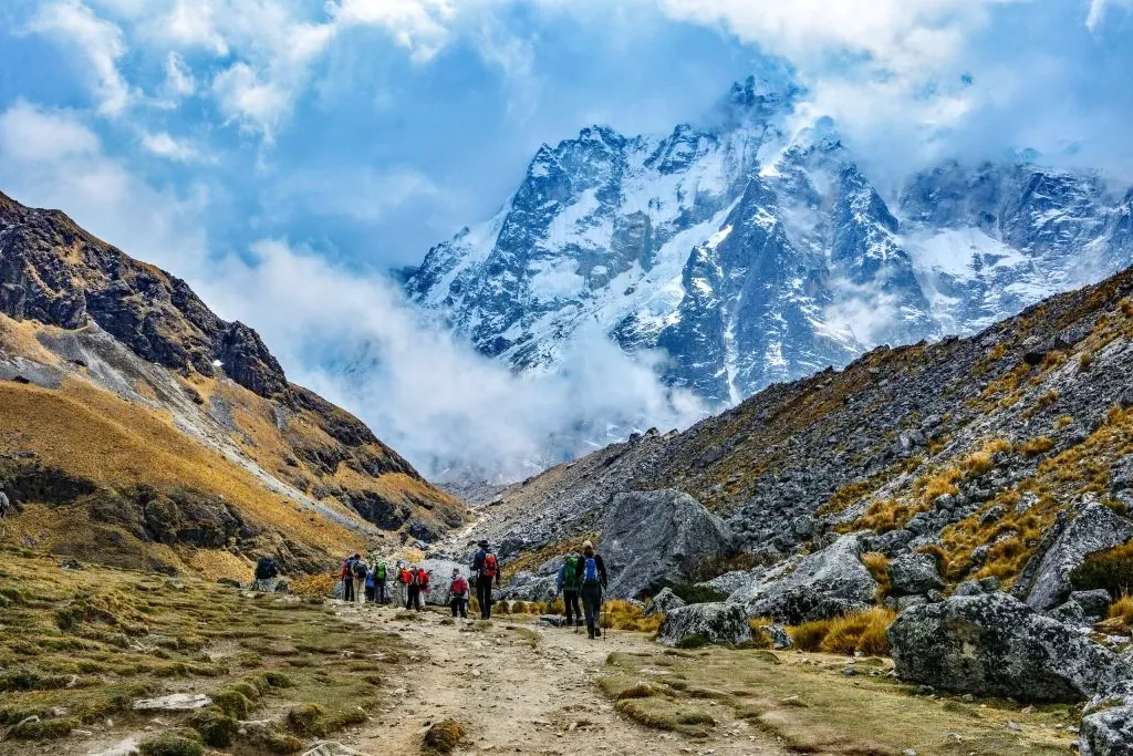 A group of trekkers walking along a high mountain path towards a majestic, cloud-covered snow peak on the Salkantay trail.
