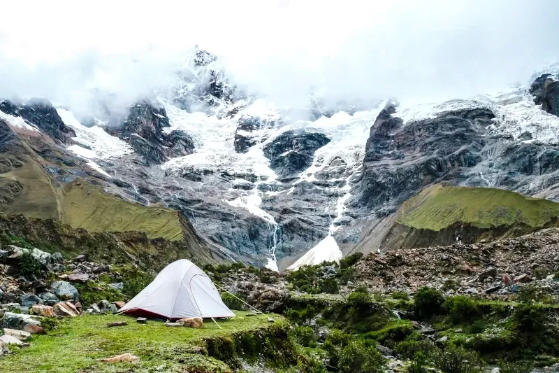 Camping at the foot of Salkantay mountain A white camping tent set up on a grassy field facing a massive snow-capped glacier mountain on the Salkantay Trek.