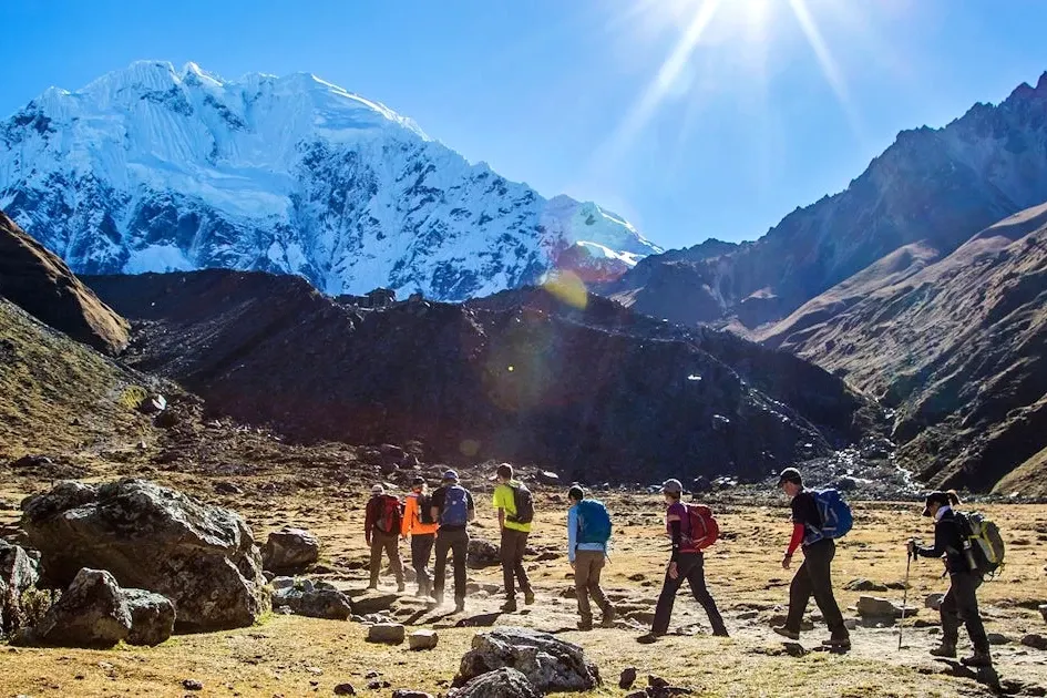 A line of hikers trekking through a wide mountain valley toward a massive white glacier under a bright sun.