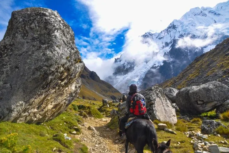 A person riding a horse on a rocky trail with the snow-capped Salkantay mountain and a blue sky in the background.